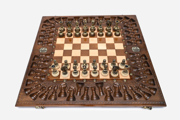 Large opened chess board with ornate bronze-and-wood chess pieces set up, framed by a deeply carved backgammon border