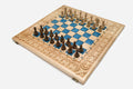 Large folding chess board in pale wood with blue epoxy squares, a carved fleur-de-lis border and wooden pieces set up for play