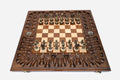 Large opened chess board with ornate bronze-and-wood chess pieces set up, framed by a deeply carved backgammon border