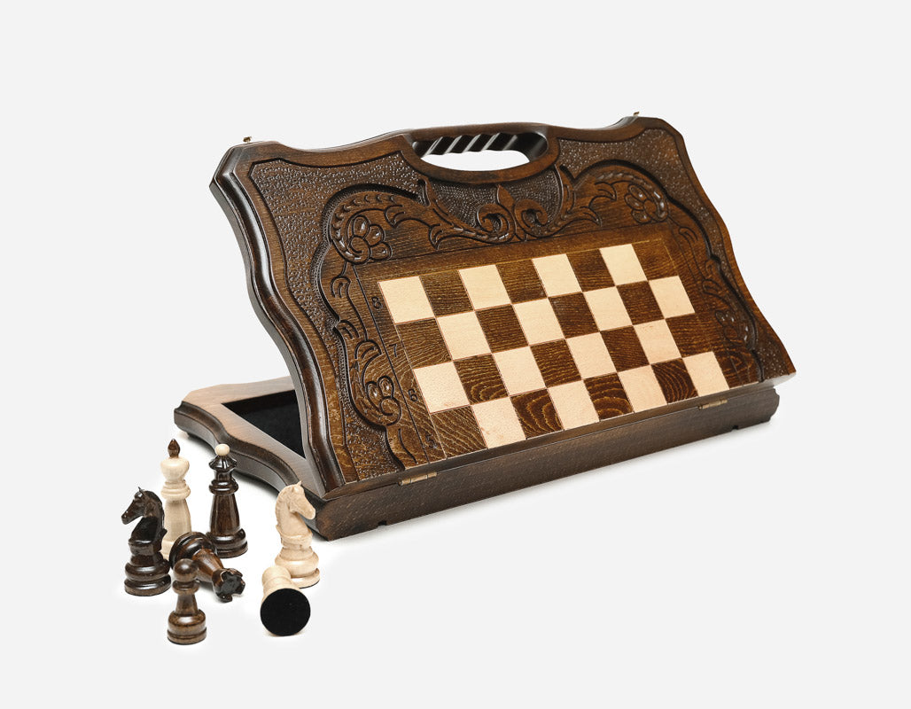 Dark walnut folding chess board standing open, showing the chessboard, floral-carved border and carry-handle cutout, with pieces in front