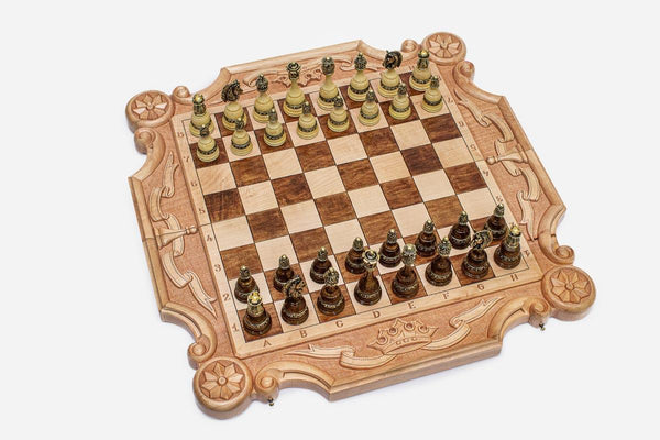 Ornately carved chess board in pale maple with floral corner medallions, ribbon scrollwork and a carved crown, set with bronze-topped wooden pieces